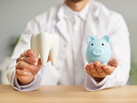 Neck-to-midsection view of man behind desk in white coat holding model tooth and blue piggy bank