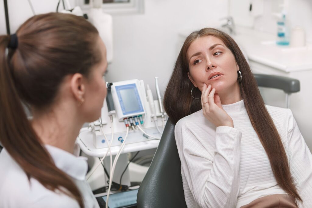 Woman in dental chair holding hand to jaw in pain looking at dentist
